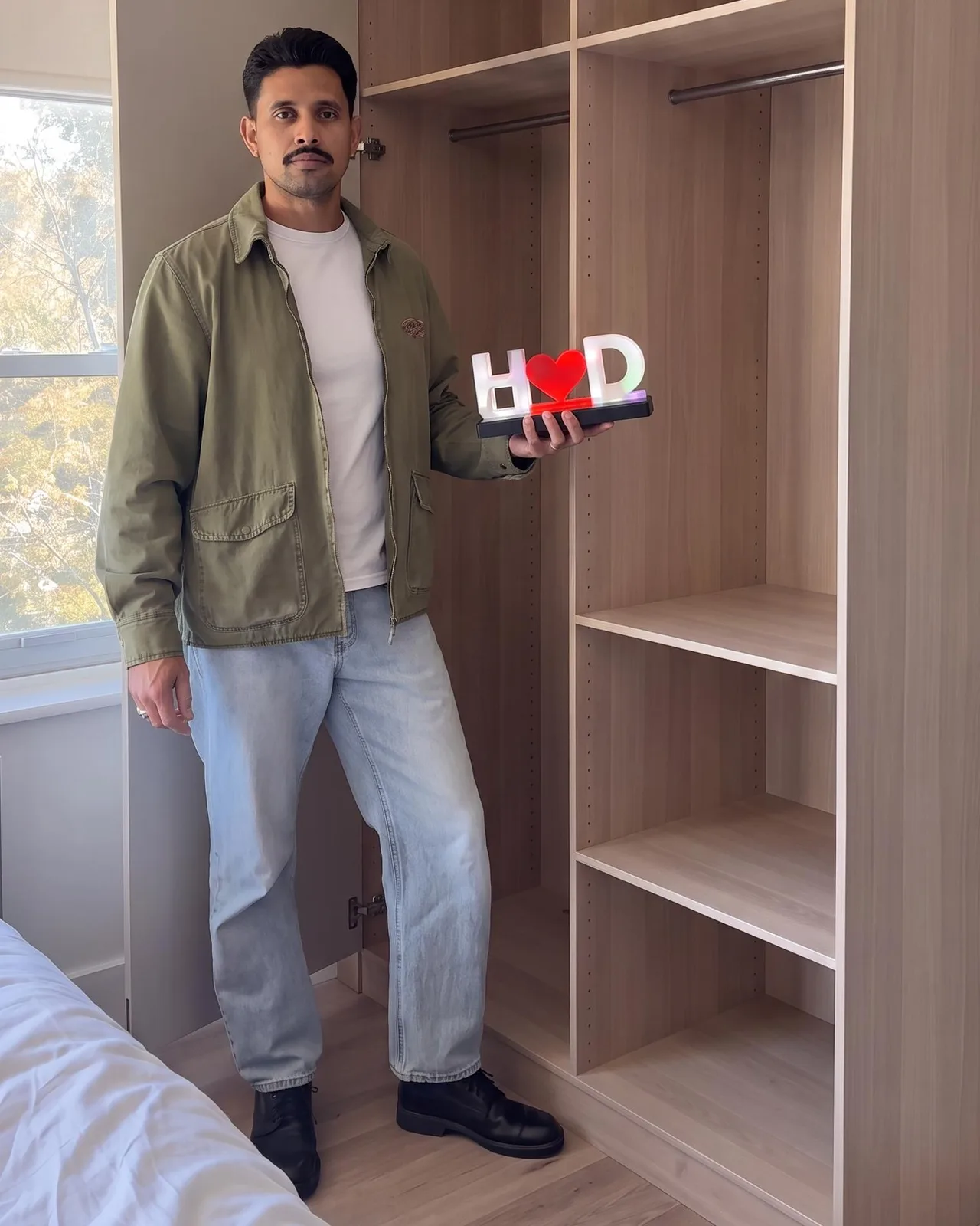 A person holding a lit H, heart, D initials name lamp in one hand beside an open wardrobe, showing the lamp’s real size