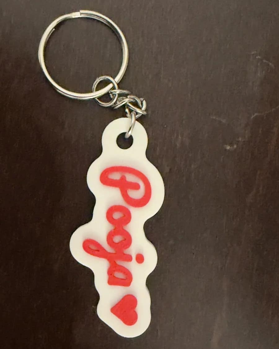 Real 3D-printed name keychain reading Pooja in red script with a heart, on a key ring, lying on a dark wooden table